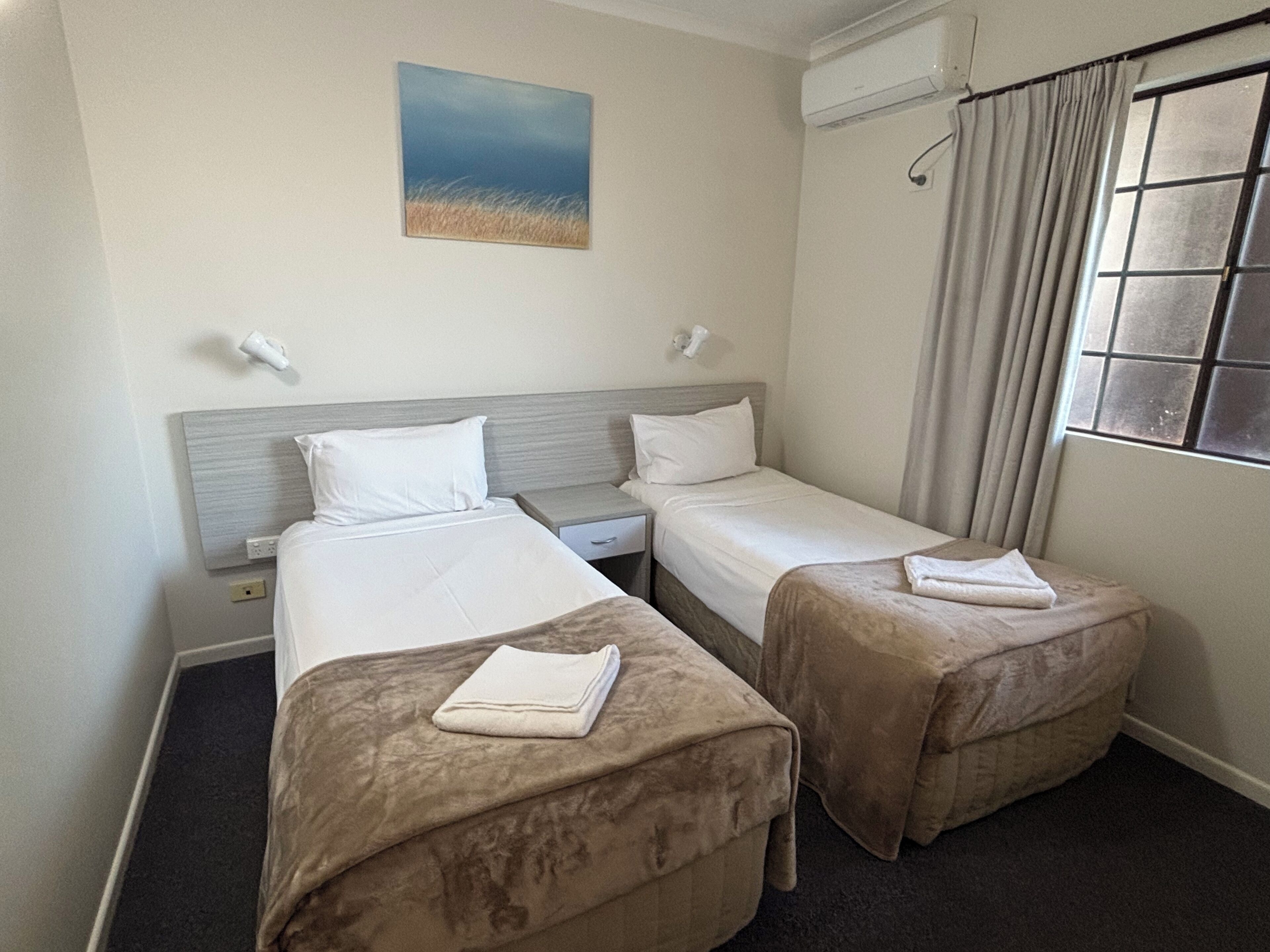 A hotel room with two beds, a painting on the wall, and an air conditioning unit.