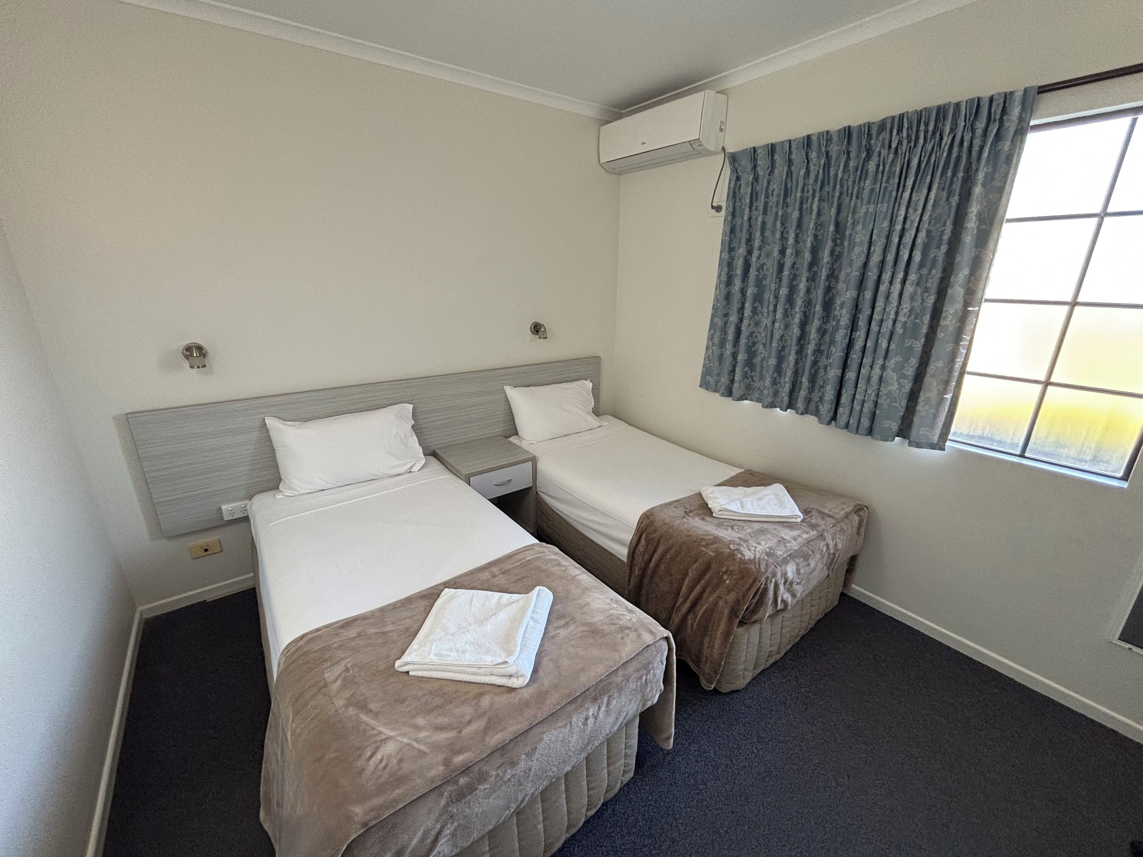 A hotel room with two beds, a window with curtains, an air conditioning unit, and a carpeted floor.