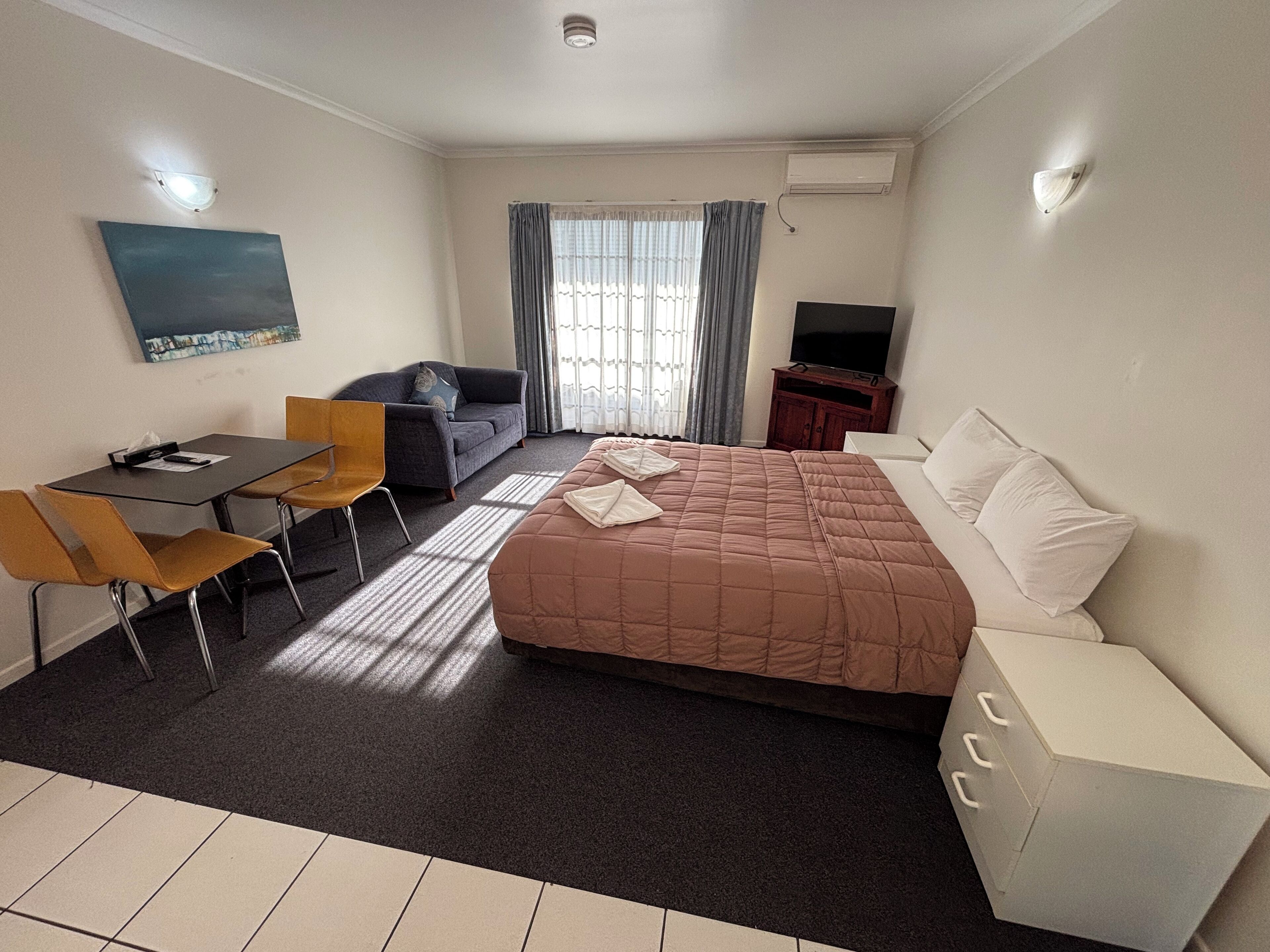 A hotel room with a bed, a sofa, a desk with chairs, a TV, and a window with curtains.