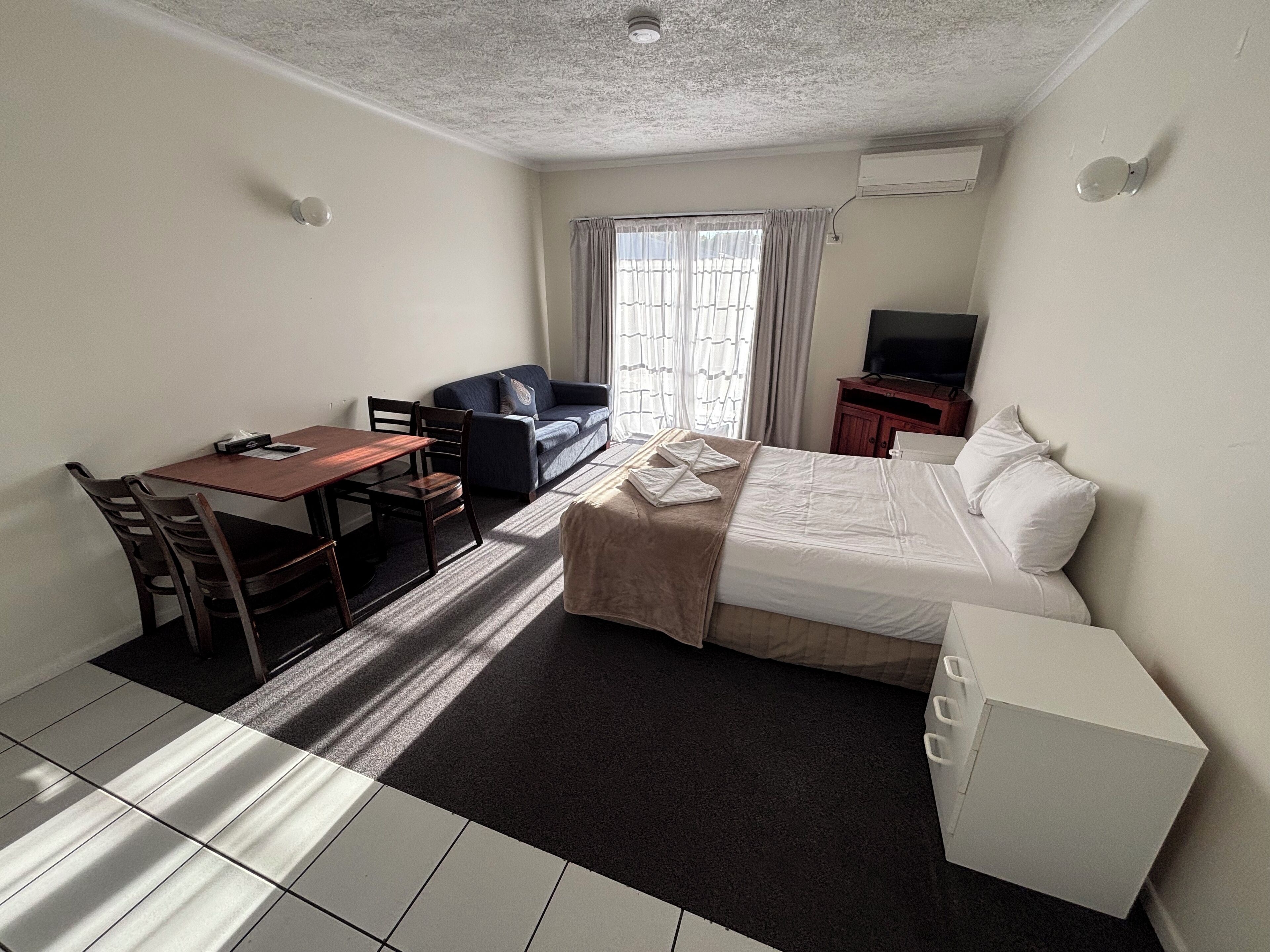 Quarto de hotel com cama, escrivaninha com cadeiras, sofá, televisão e pequeno armário.