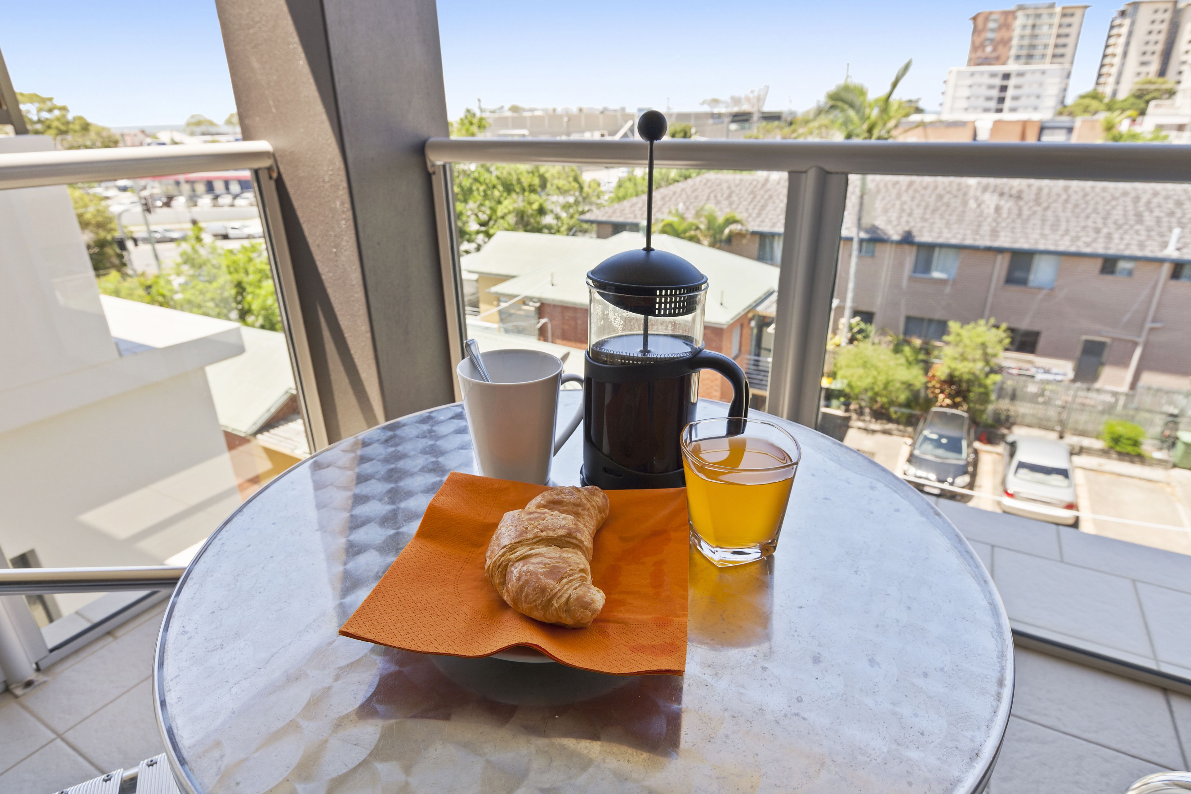 A balcony with a table, a coffee maker, a cup, a glass of tea, and a croissant.