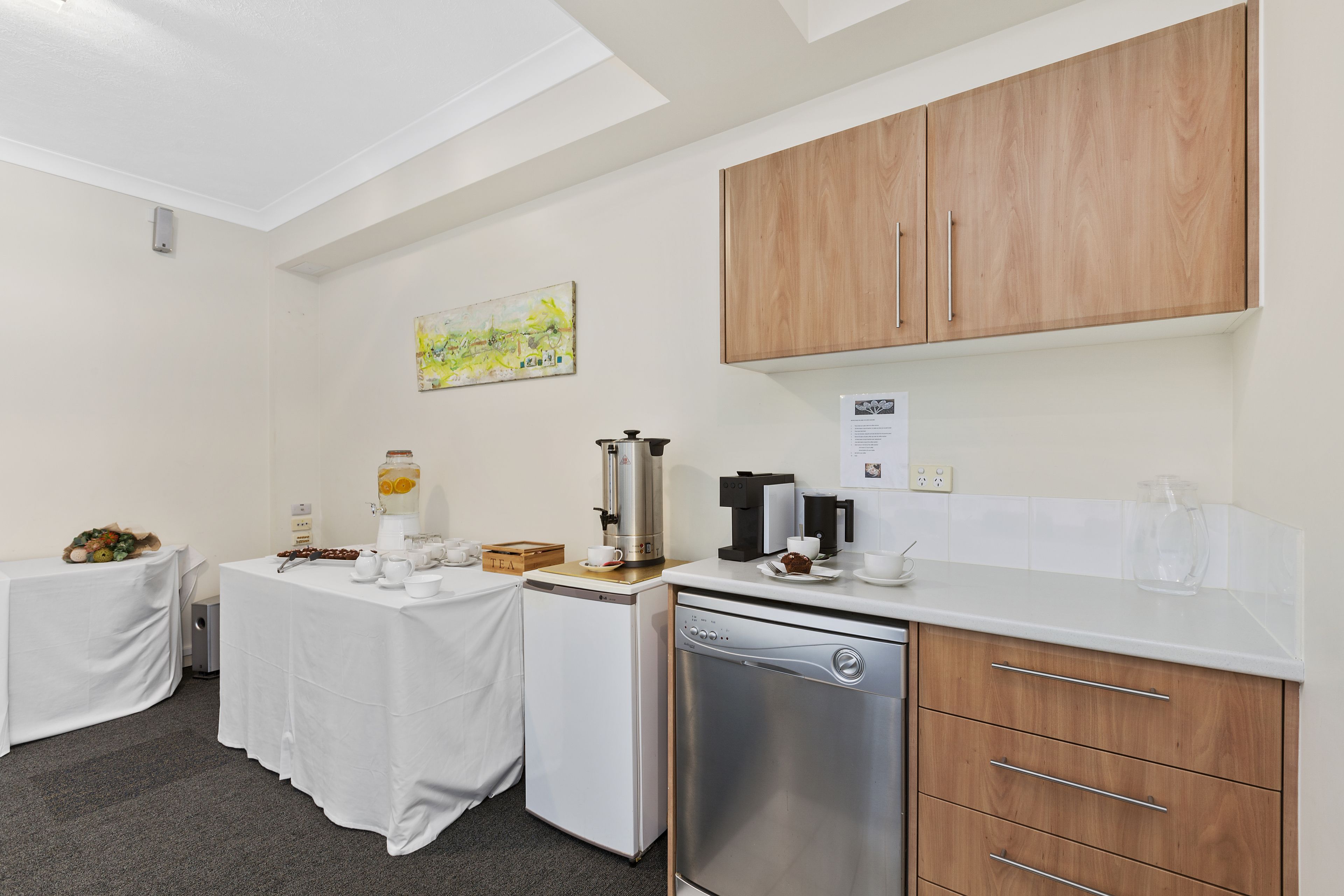A compact kitchen with a coffee machine, a dishwasher, and a small refrigerator. There is a table with a fruit basket and a coffee pot.