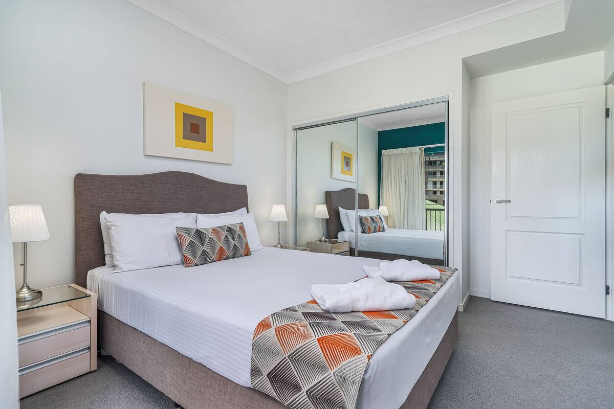 A hotel room with a large bed, a bedside table with lamps, a mirror, and a wardrobe.