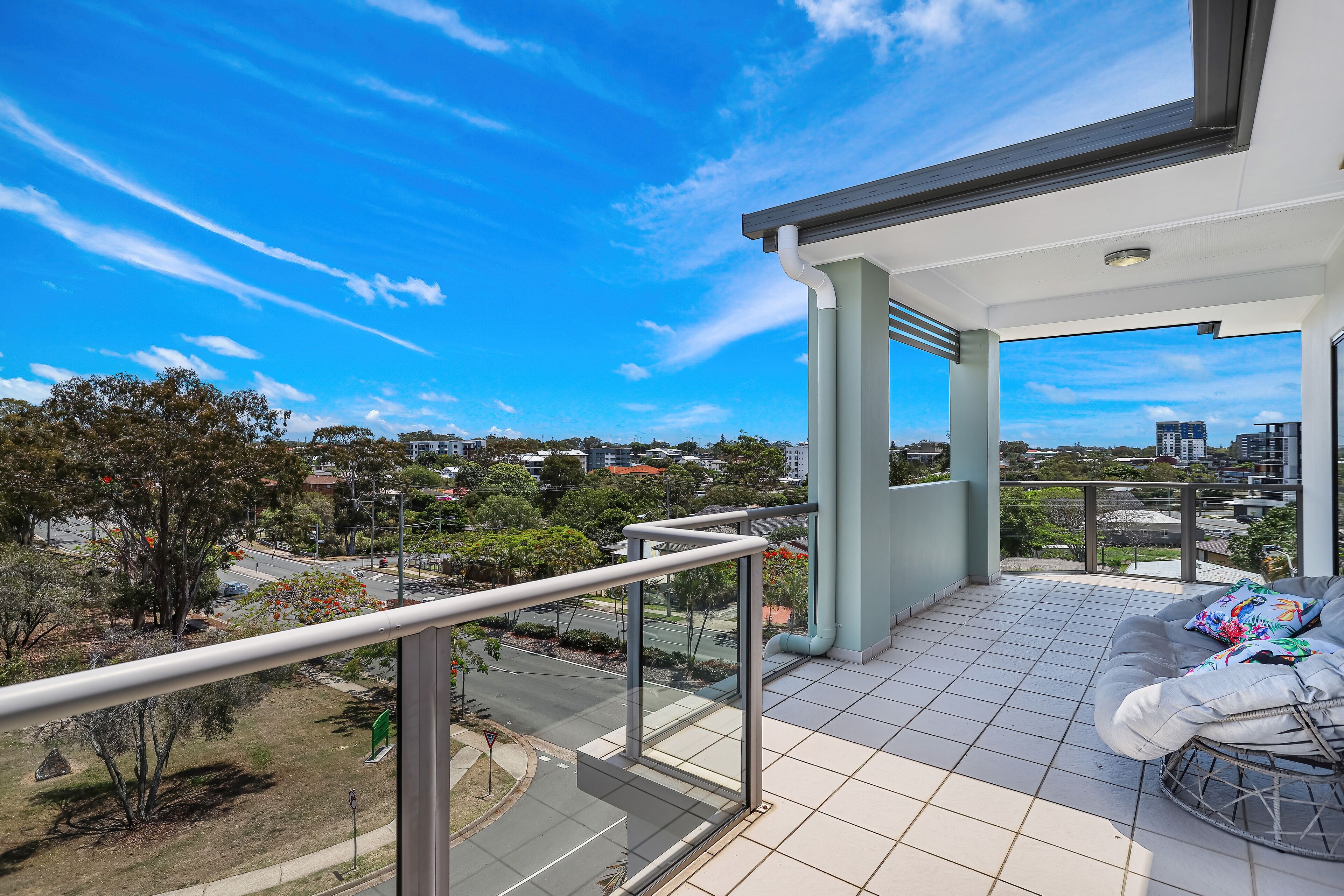 A balcony with a glass railing, a cushioned seating area, and a city view.
