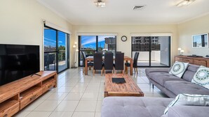 A spacious living room with a large flat-screen TV, a grey sofa, a wooden coffee table, and a dining area with a table and chairs. - La Vida on Anzac (Redcliffe)