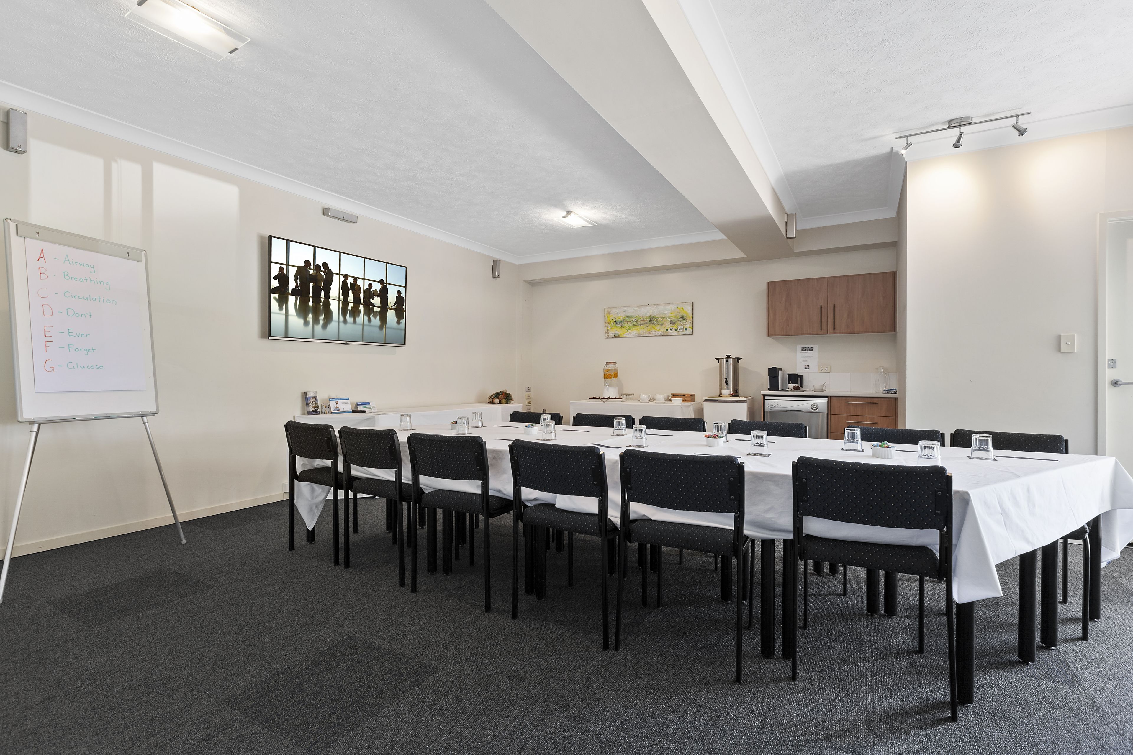 Una sala de conferencias con una pizarra, mesas, sillas y una zona de cocina al fondo.