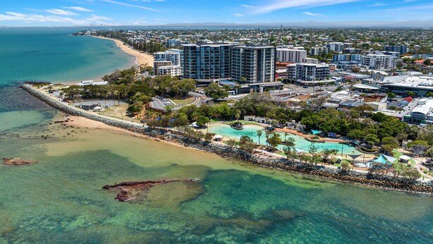 An aerial view of a coastal resort with high-rise buildings, a beach, and a clear blue sky. - La Vida on Anzac (Redcliffe)