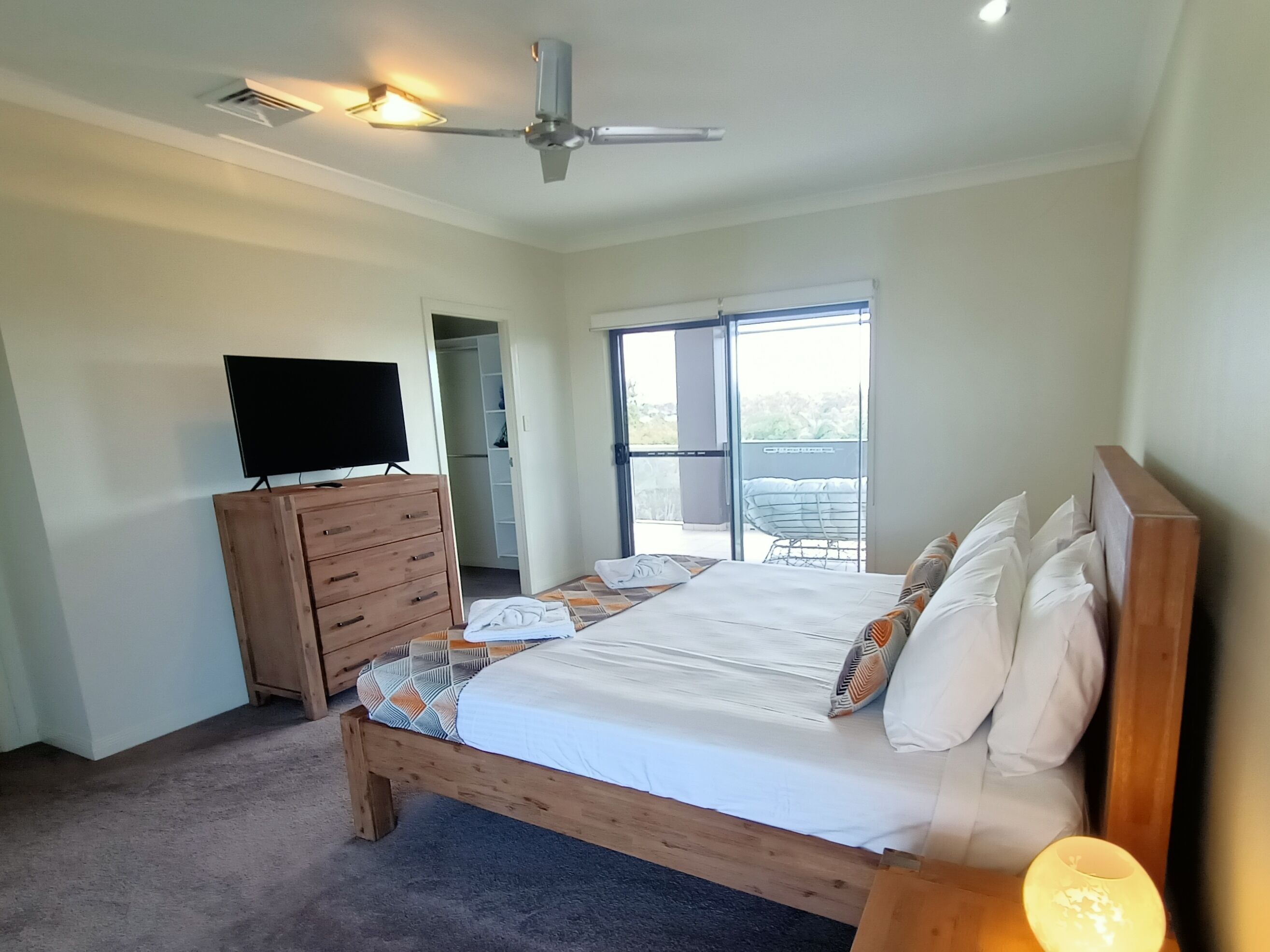 Quarto com cama, penteadeira de madeira, televisão, ventoinha no teto e varanda com cadeira.