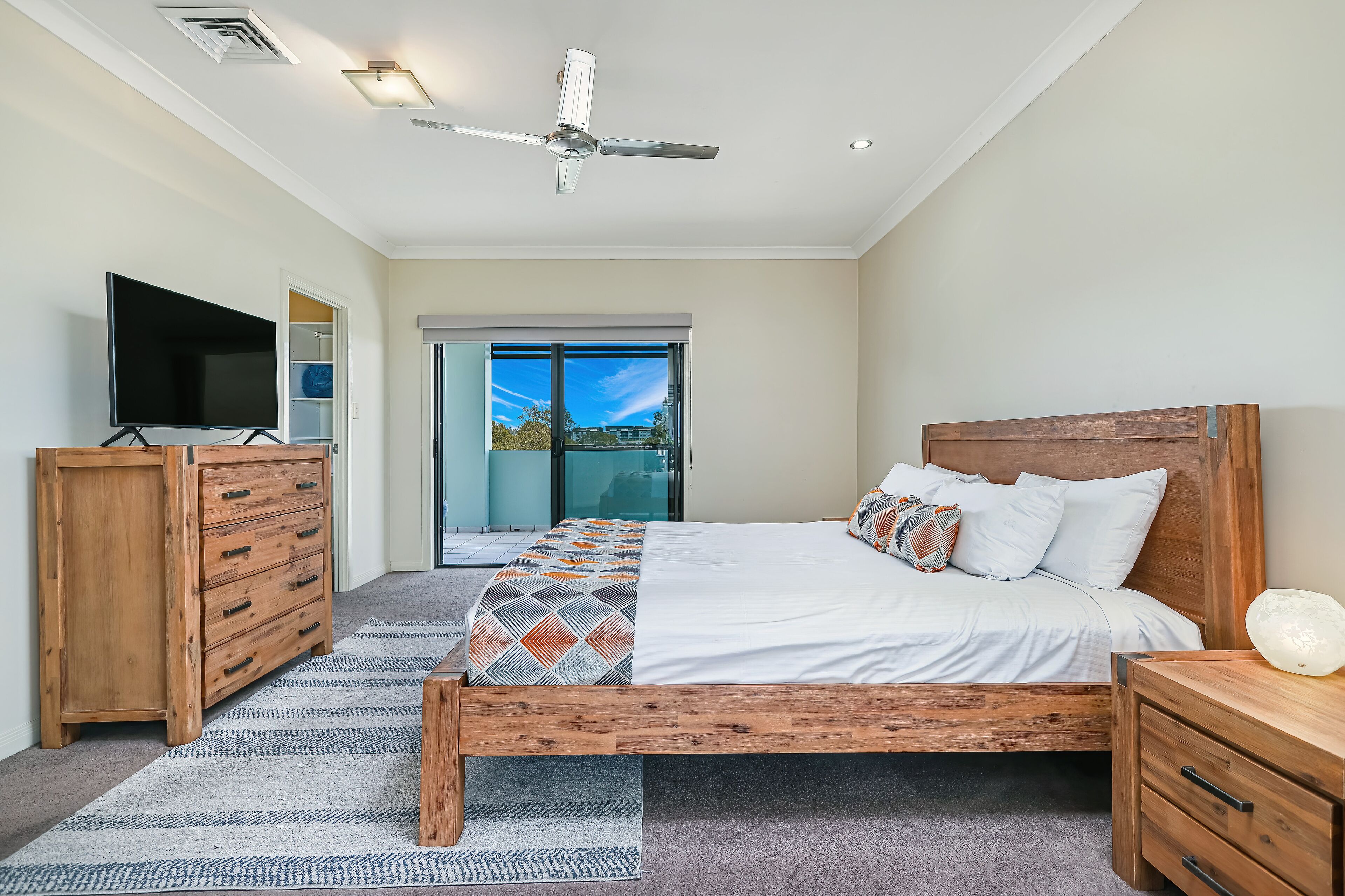 A modern bedroom with a wooden bed, a TV, a ceiling fan, and a view of the outdoors through a glass door.