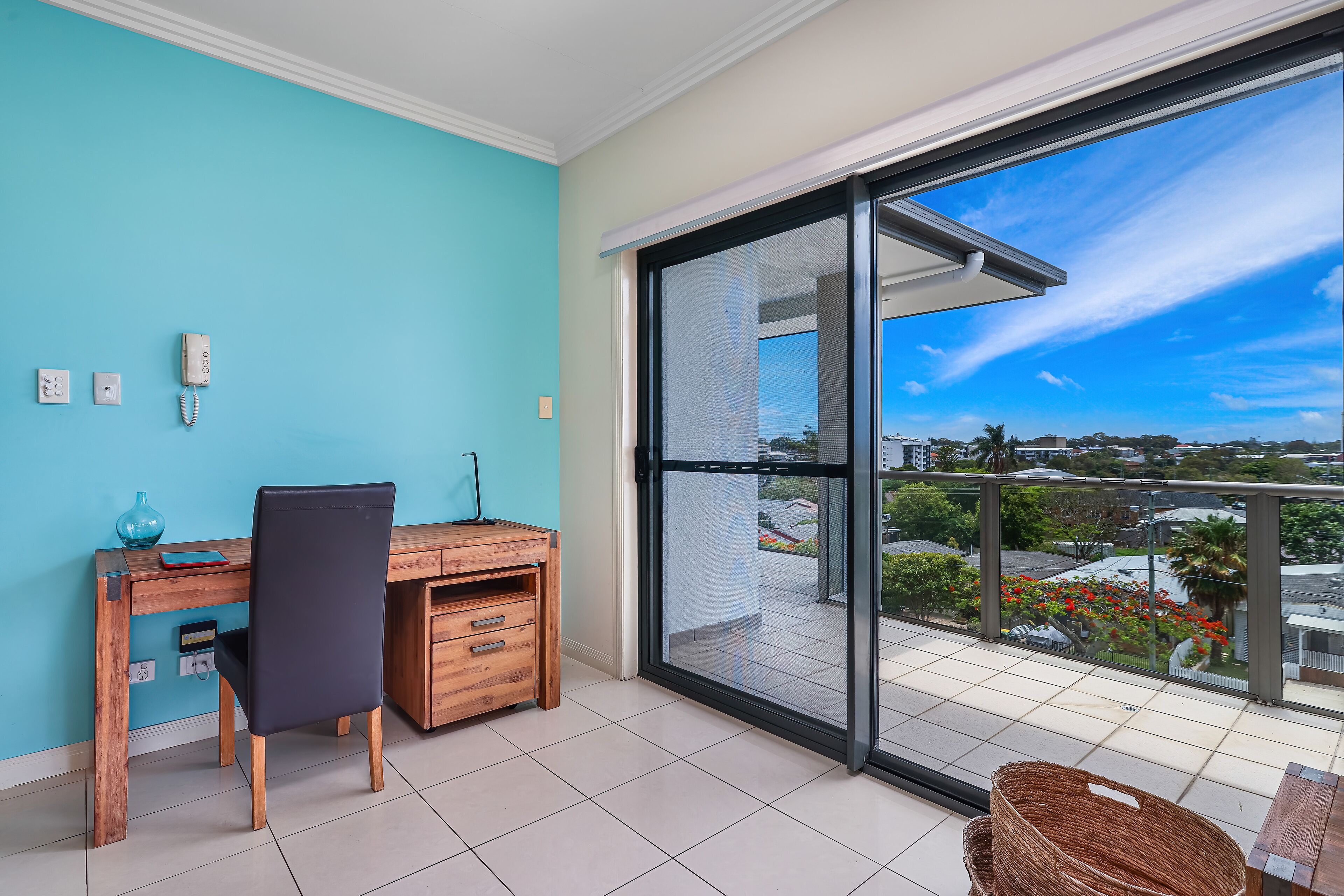 A room with a wooden desk, a chair, a large sliding glass door to a balcony, and a view of a cityscape.