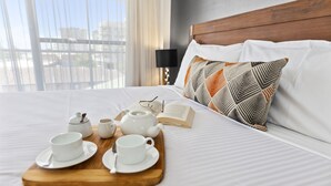 A neatly made bed with a wooden tray holding cups and a teapot, an open book, and a pillow with a geometric pattern. - La Vida on Anzac (Redcliffe)
