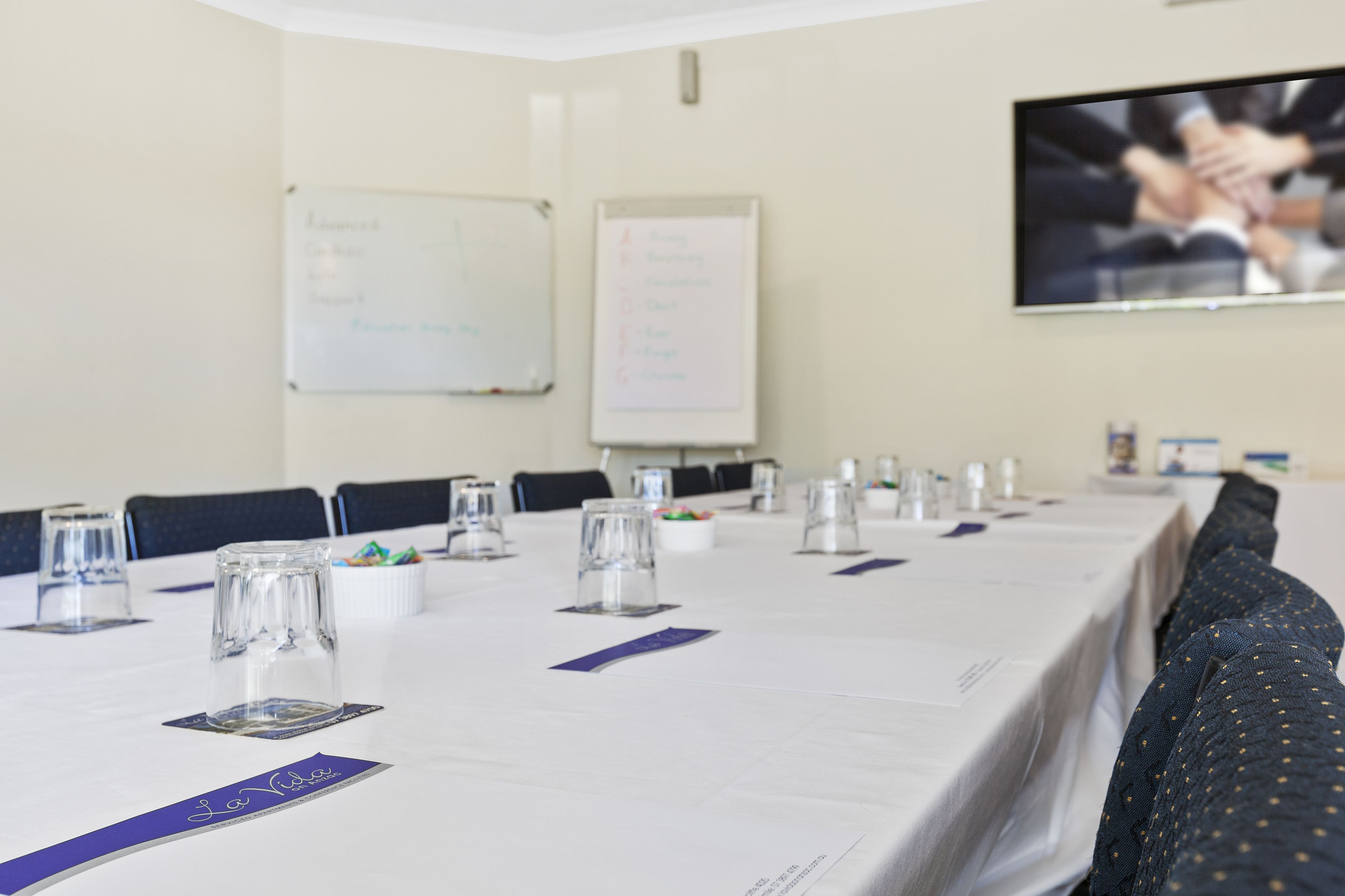 A conference room with a long table set for a meeting, whiteboards on the walls, and a television screen displaying a group of people.