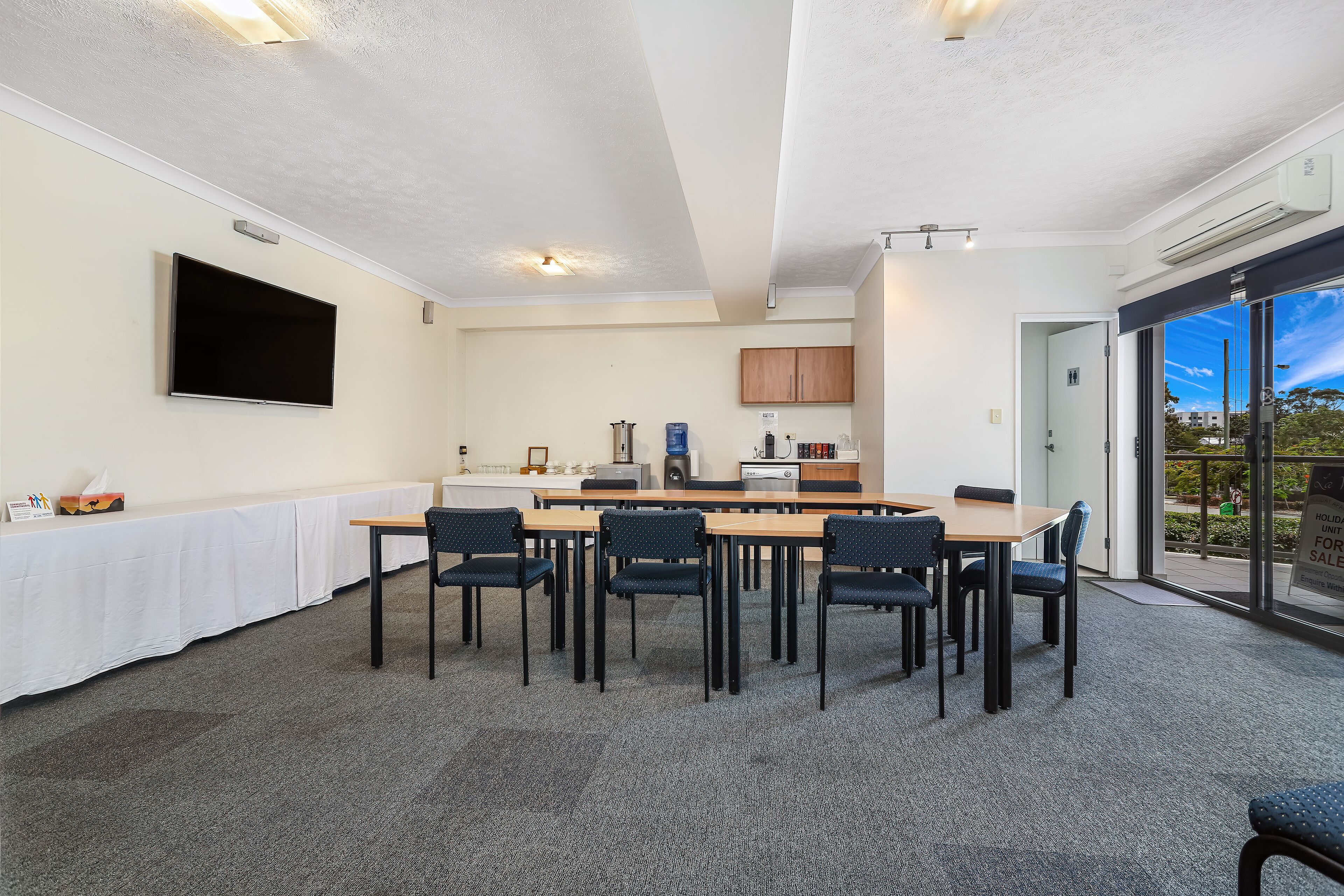 A conference room with a long table, chairs, a flat-screen TV, and a kitchen area with cabinets and appliances.