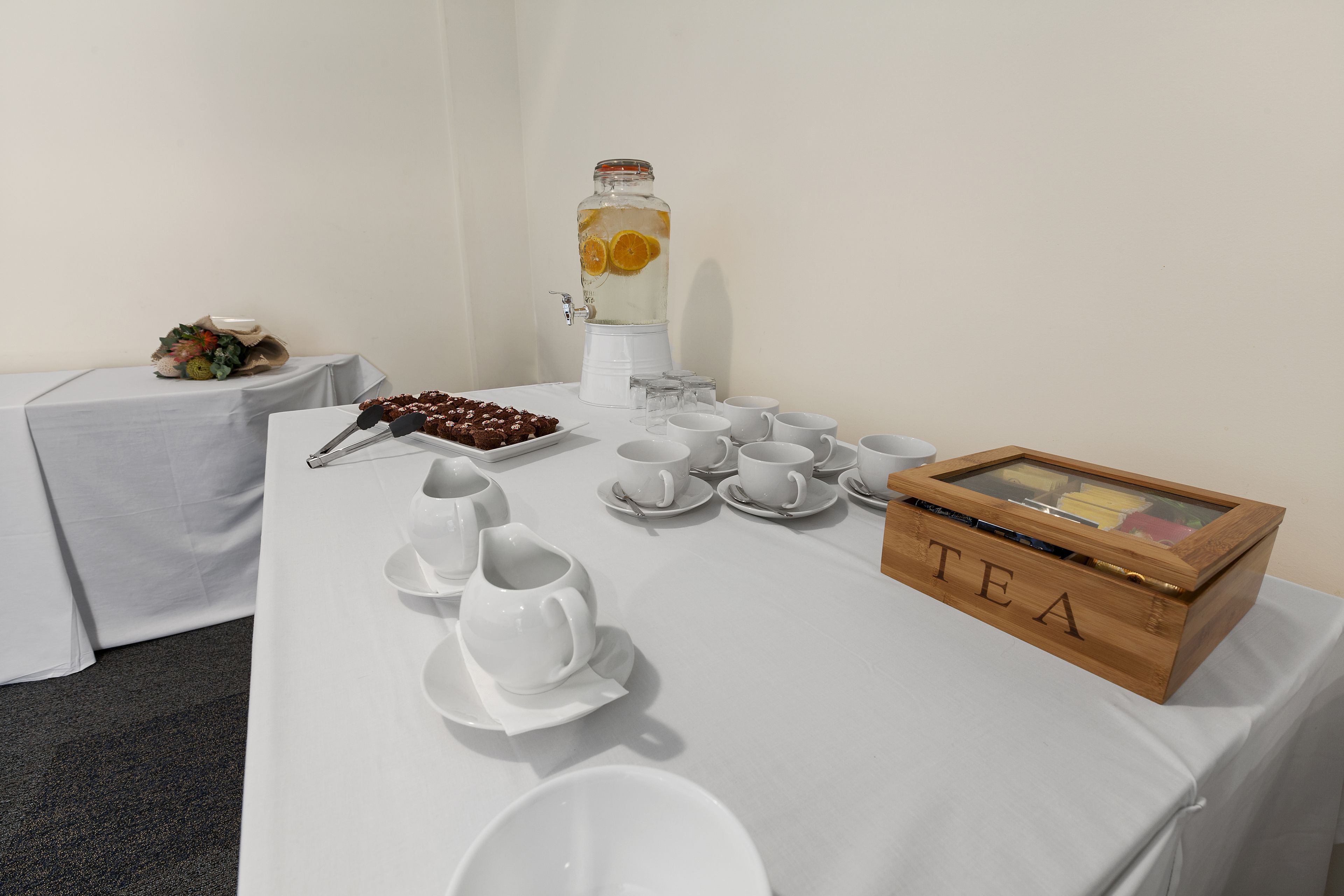 Une table dressée avec une vaisselle à thé blanche, un grand pichet rempli de tranches de fruits et une boîte en bois étiquetée « THÉ ».
