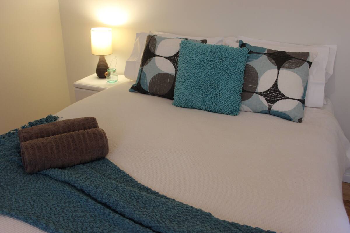 A neatly made bed with a blue blanket and two brown towels, a bedside lamp, and patterned pillows.