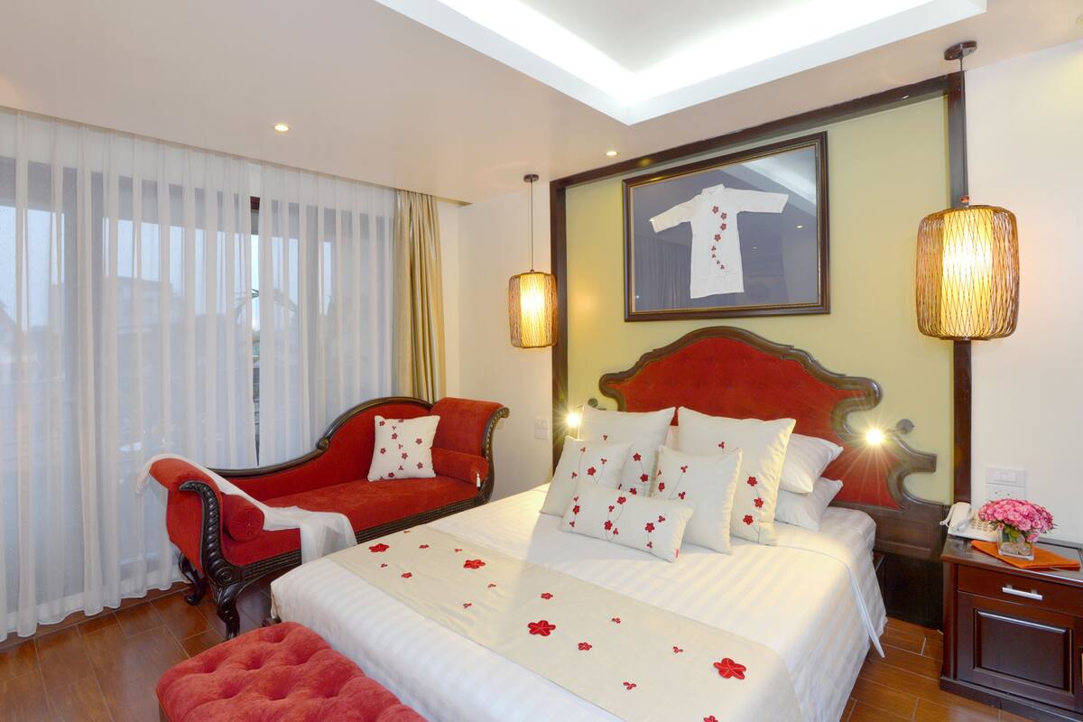 A hotel room with a large bed, a red armchair, a wooden nightstand, and a large window with sheer curtains.