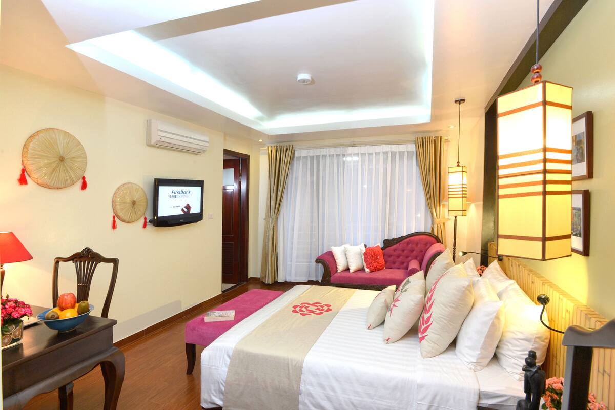 A hotel room with a bed, a TV, a dining table with fruit, and a window with curtains.