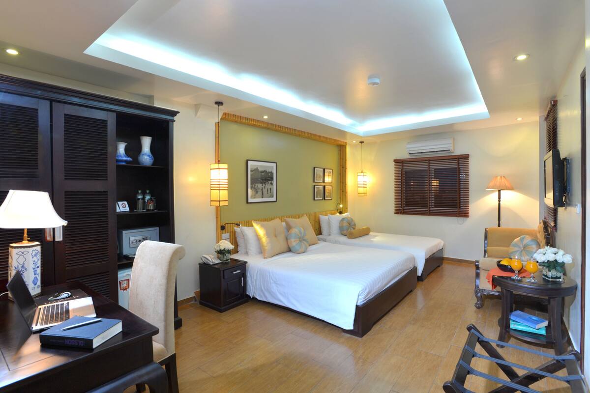 A hotel room with two beds, a desk with a laptop, a chair, a small table with a lamp, and a TV.