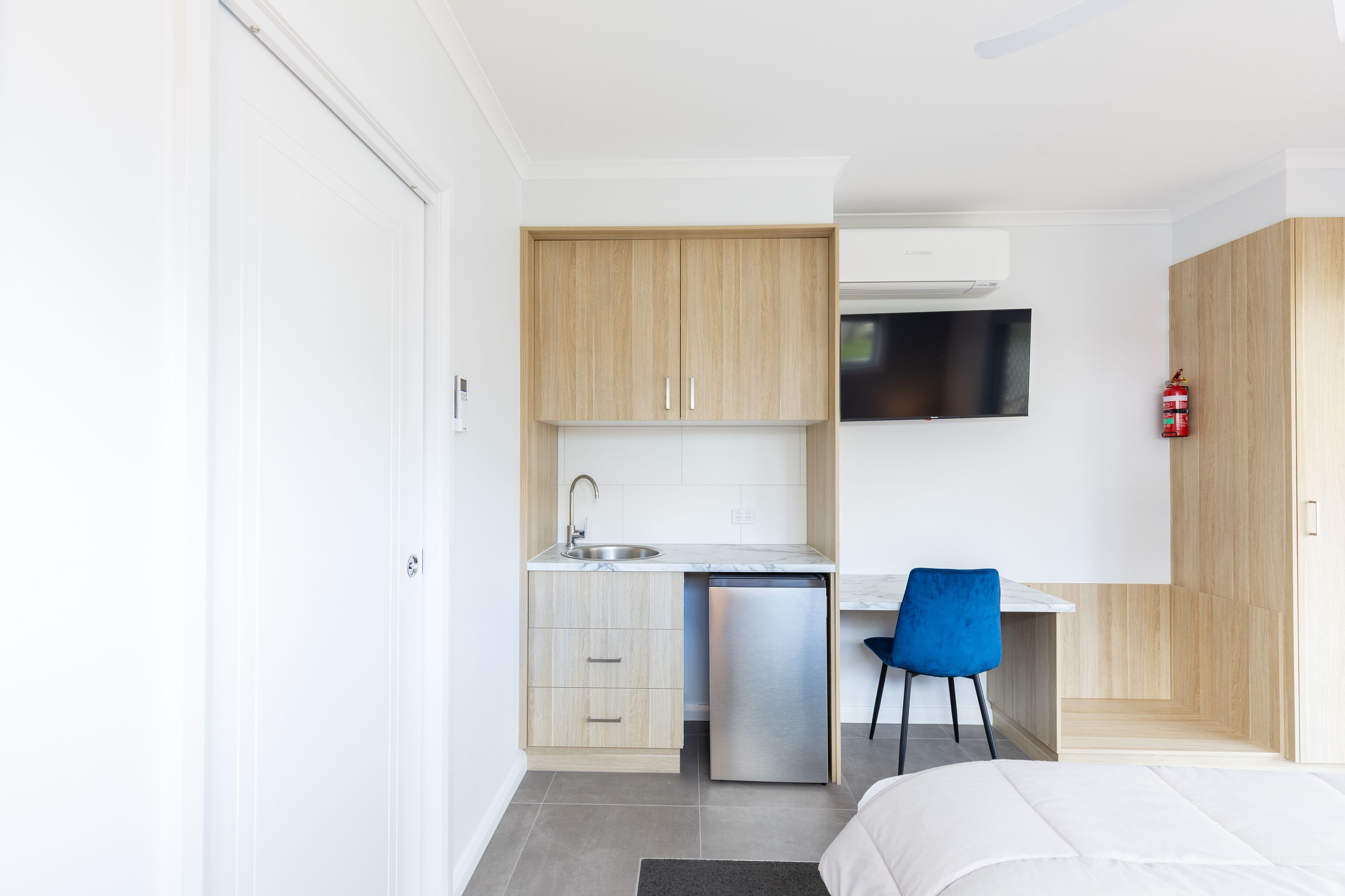 A compact hotel room with a kitchenette, a bed, a blue chair, and a mounted television.