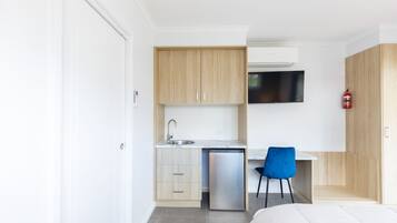 A compact hotel room with a kitchenette, a bed, a blue chair, and a mounted television.