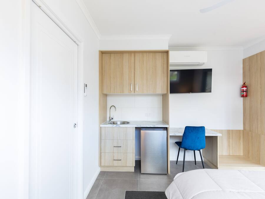 A compact hotel room with a kitchenette, a bed, a blue chair, and a mounted television.