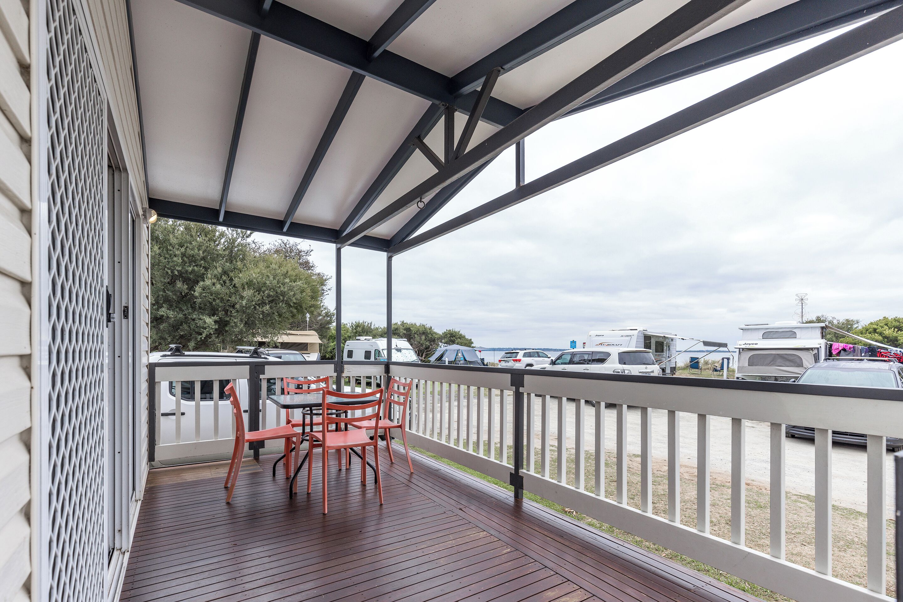 A covered outdoor area with a wooden deck, a table with chairs, and a view of parked vehicles and trees in the background.