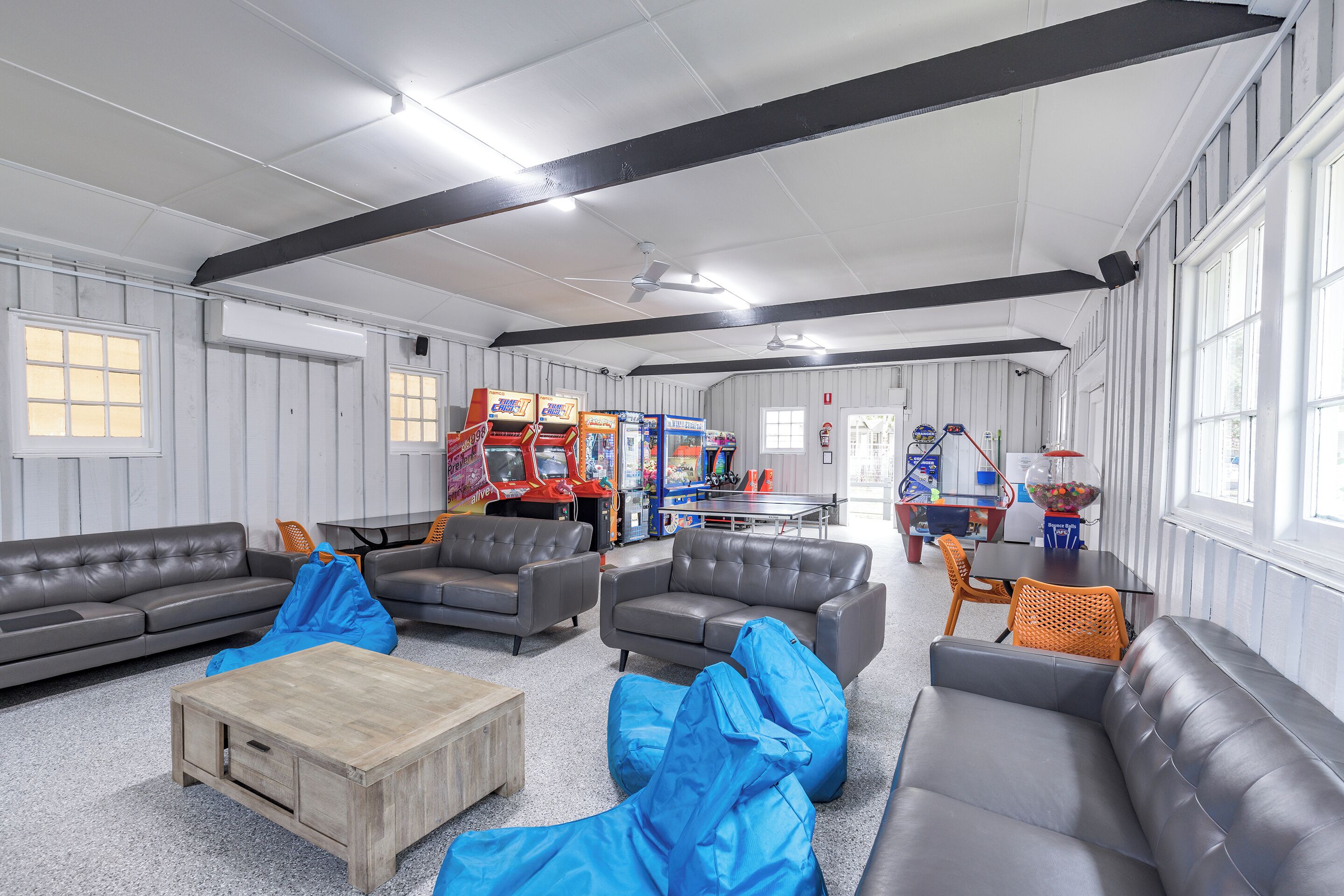 A spacious room with grey leather sofas, a wooden coffee table, and blue bean bags. There are arcade games and a ping pong table in the background.