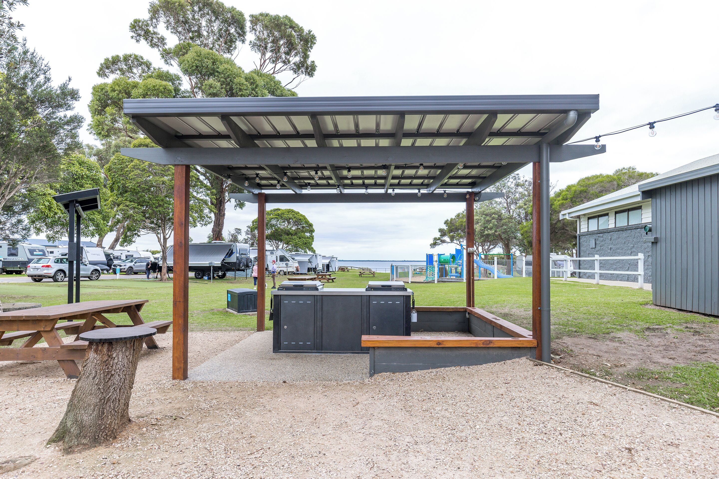 Un área de estar al aire libre cubierta con una mesa y bancos, rodeada de árboles y un parque infantil al fondo.