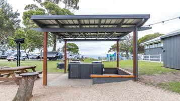 Un área de estar al aire libre cubierta con una mesa y bancos, rodeada de árboles y un parque infantil al fondo.