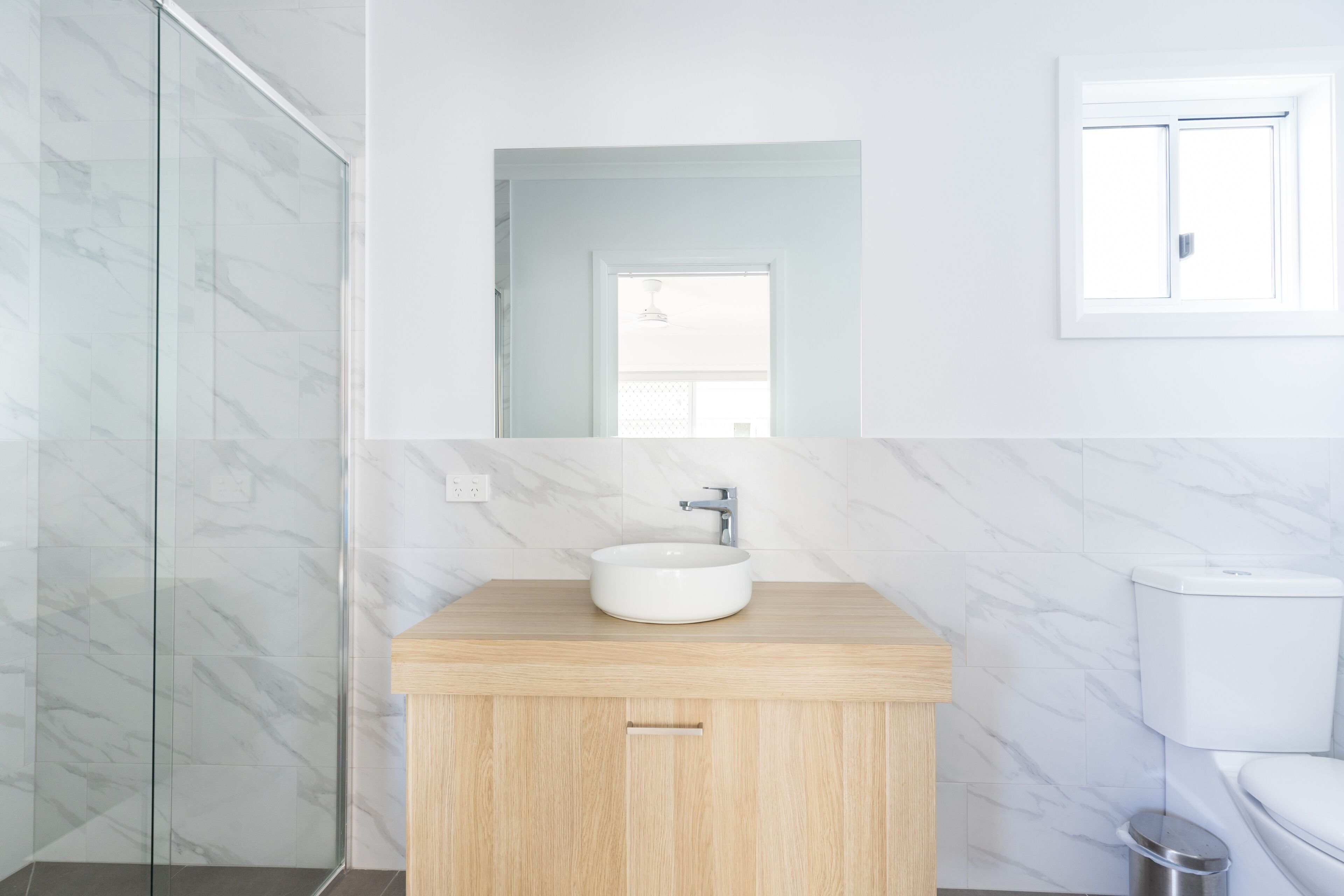 Un baño con pared de mármol, tocador de madera, lavabo blanco y retrete.