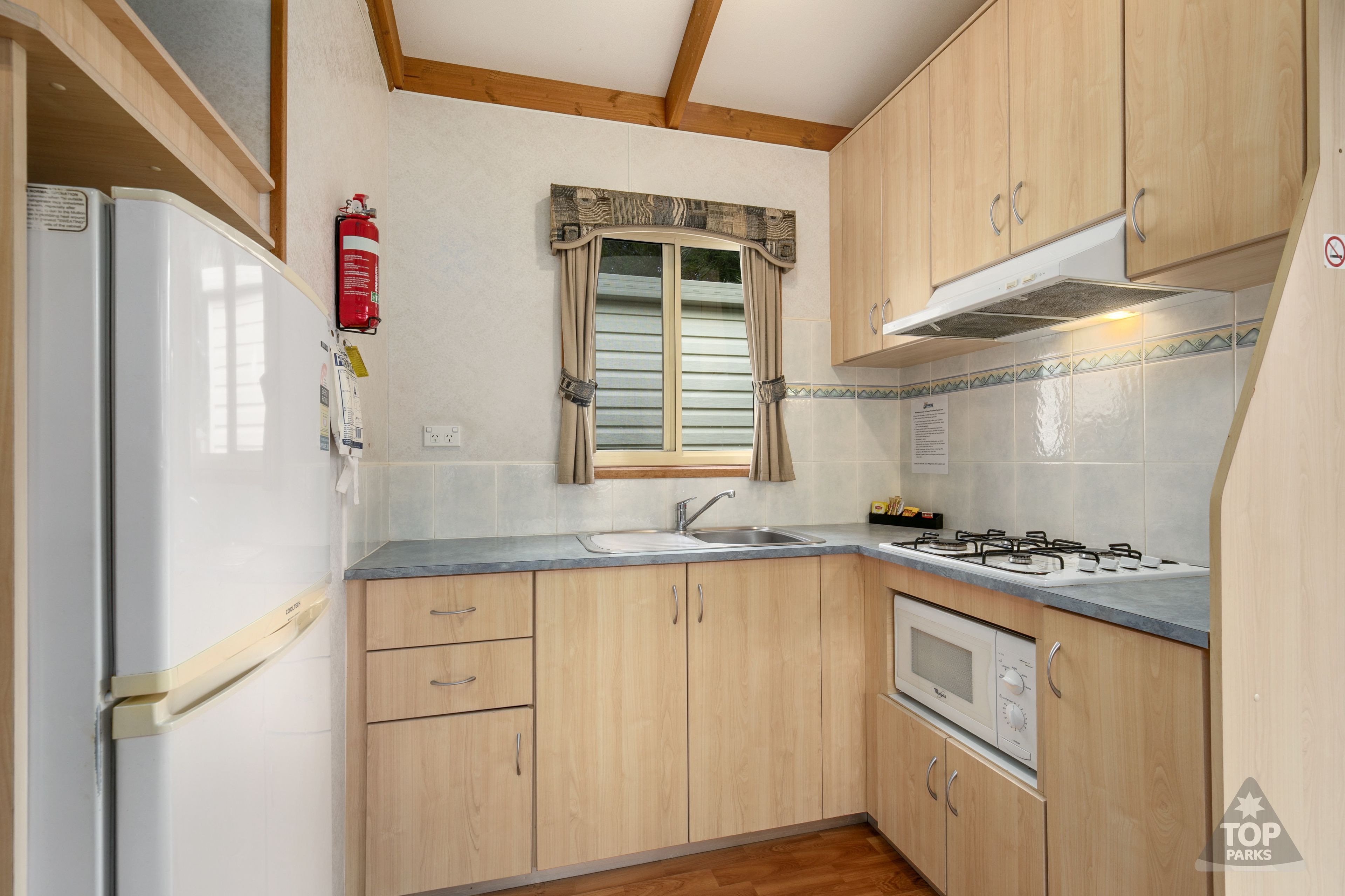 A compact kitchen with wooden cabinets, a built-in microwave, a gas stove, and a window with curtains.