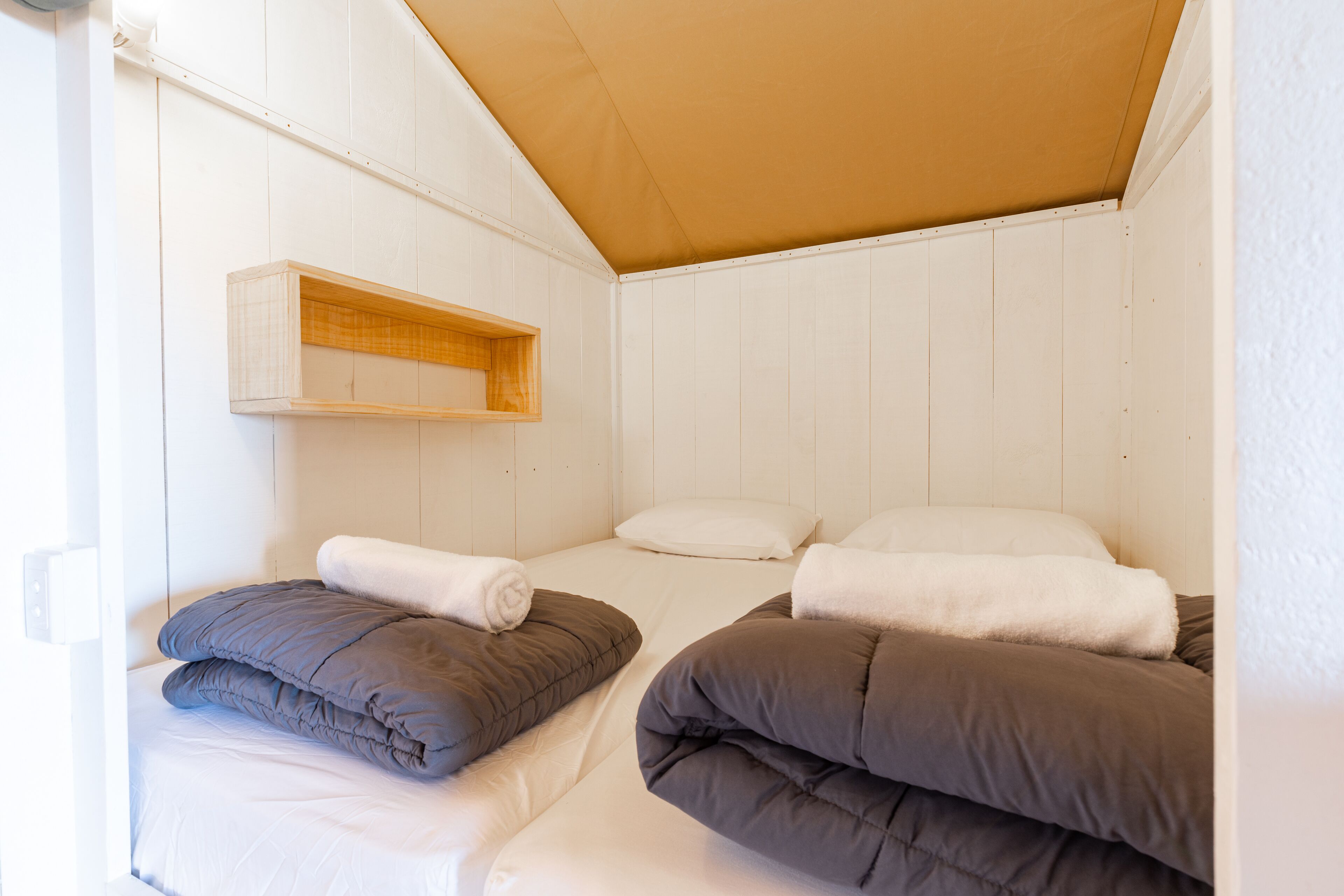 A small, cozy room with two beds, each with a pillow and a rolled-up towel. There is a wooden shelf above the beds.