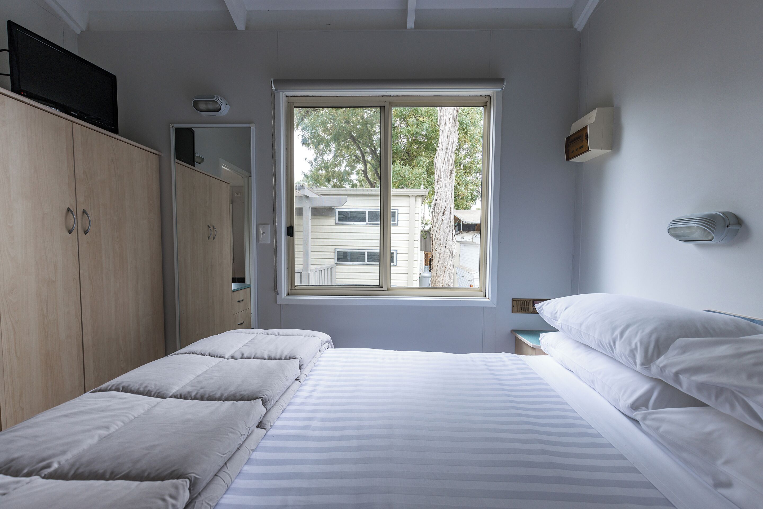 A bedroom with a large window, a bed, a wardrobe, and a bedside table.