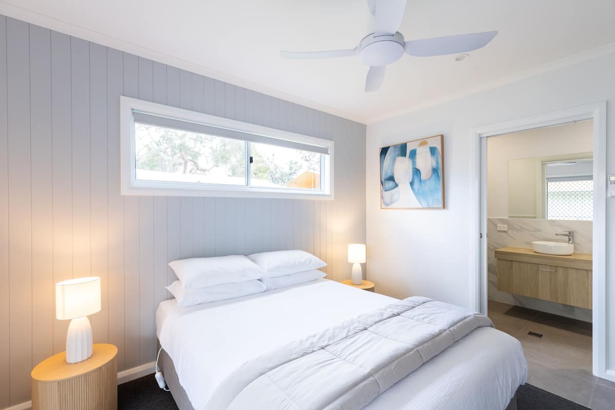 Une chambre à coucher avec un lit, deux lampes de chevet, un ventilateur de plafond, une fenêtre et une salle de bain visible à travers une porte ouverte.