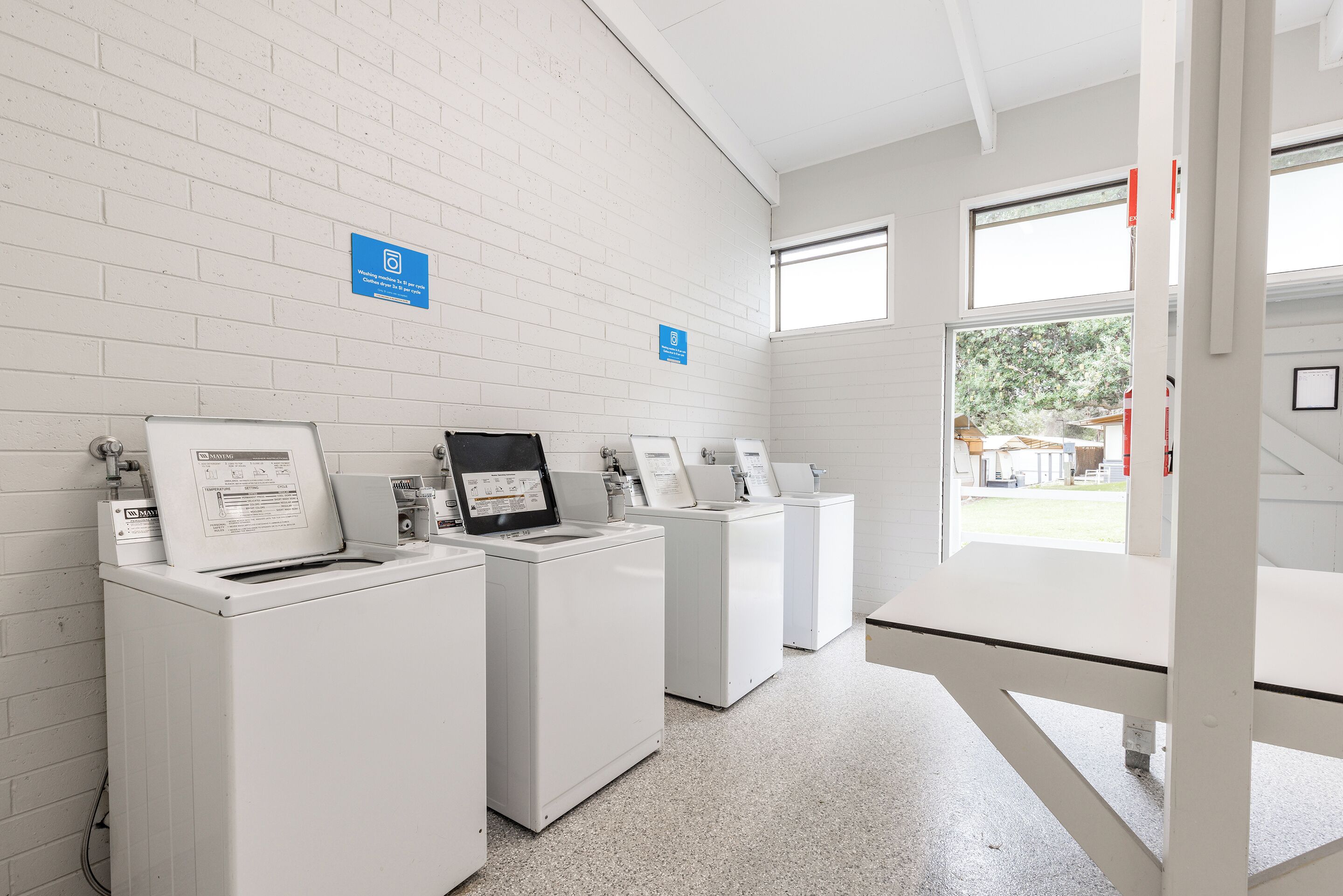 A laundry facility with four washing machines, a white brick wall, and a window providing natural light.