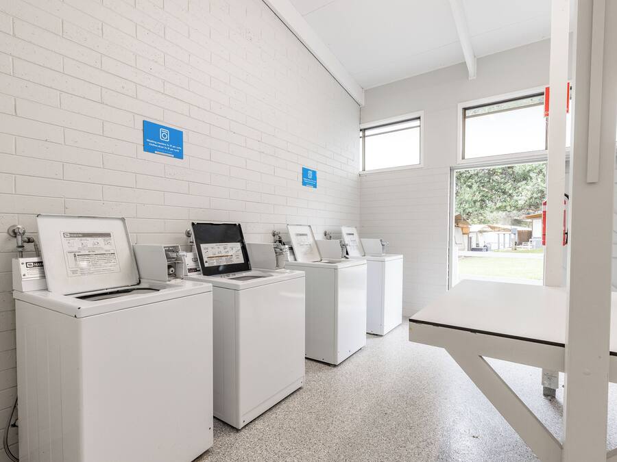 A laundry facility with four washing machines, a white brick wall, and a window providing natural light.