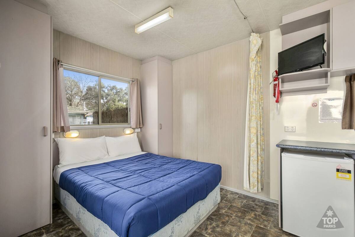 A compact room with a bed, a small kitchenette, a television, and a window with curtains.