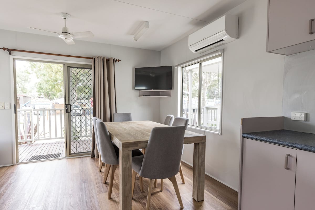 Une cuisine moderne dotée d’une cuisinière intégrée, d’un coin repas avec une table en bois et des chaises grises, et d’une grande fenêtre qui laisse entrer la lumière naturelle.