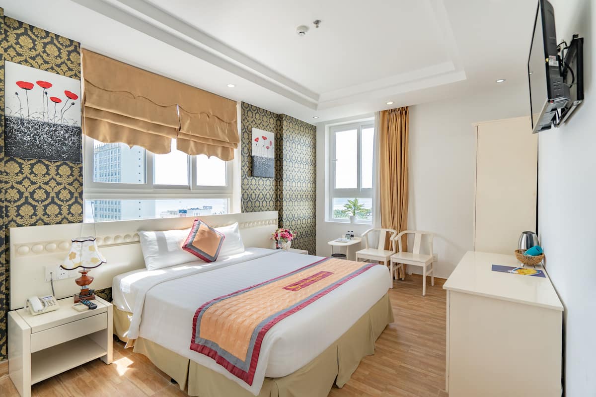 A hotel room with a large bed, bedside table, lamp, and a desk with a chair.
