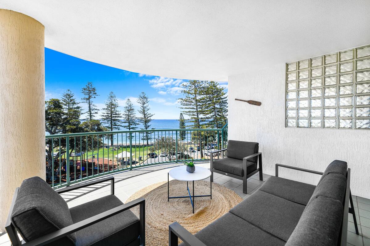 Een balkon met uitzicht op zee, een ronde tafel met een plant, en grijze banken.