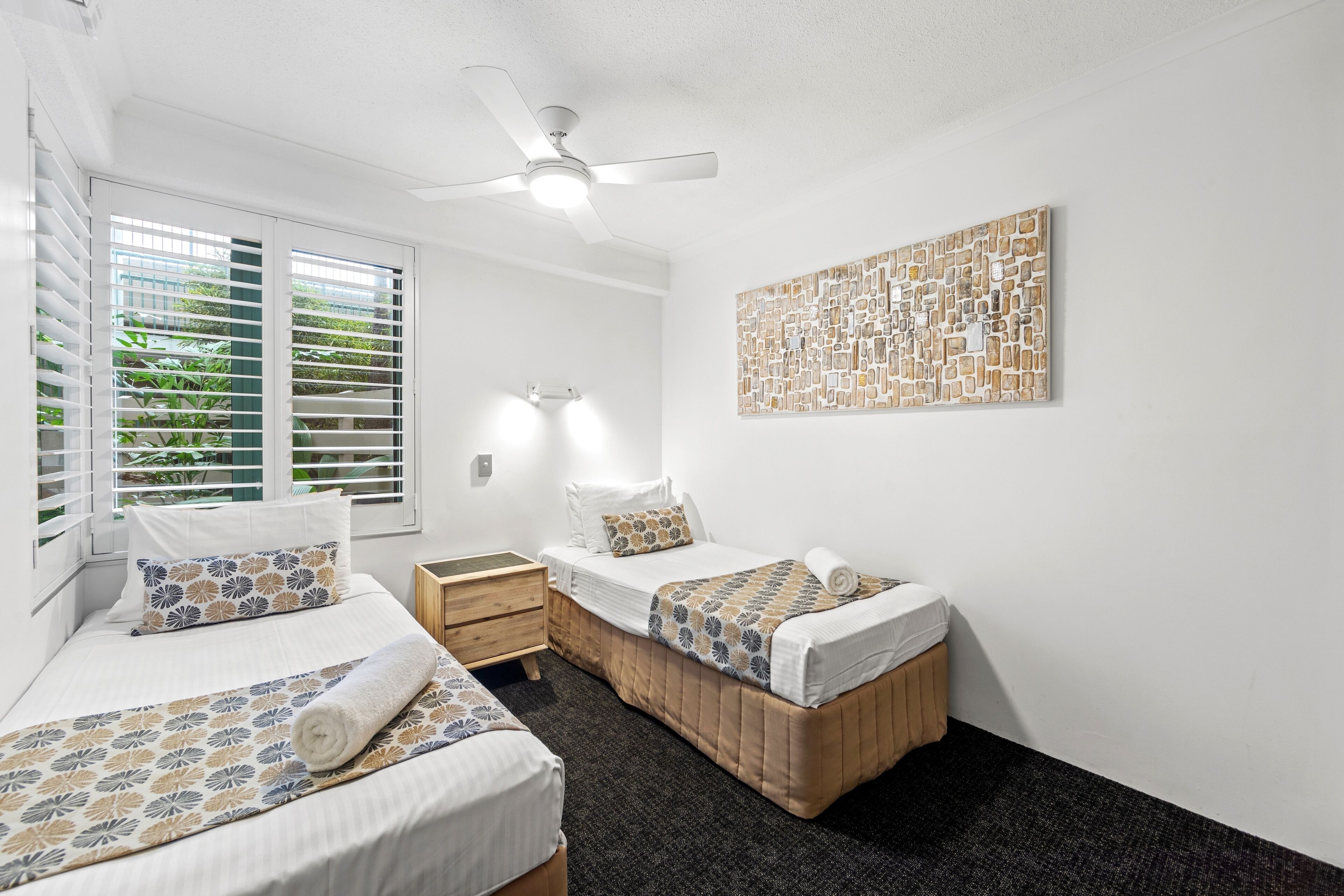 Un dormitorio con dos camas, un ventilador de techo, una obra de arte en la pared y una ventana con persianas.