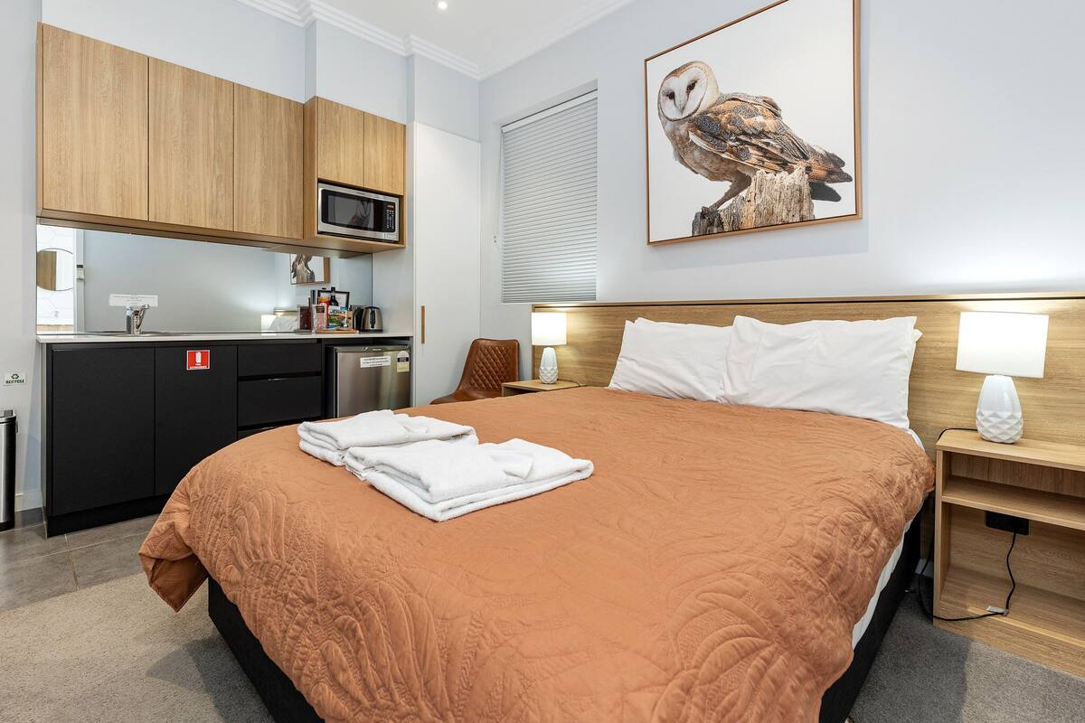 A hotel room with a large bed, a wooden headboard, a wall-mounted TV, a microwave, a small kitchen area, and a framed owl picture.