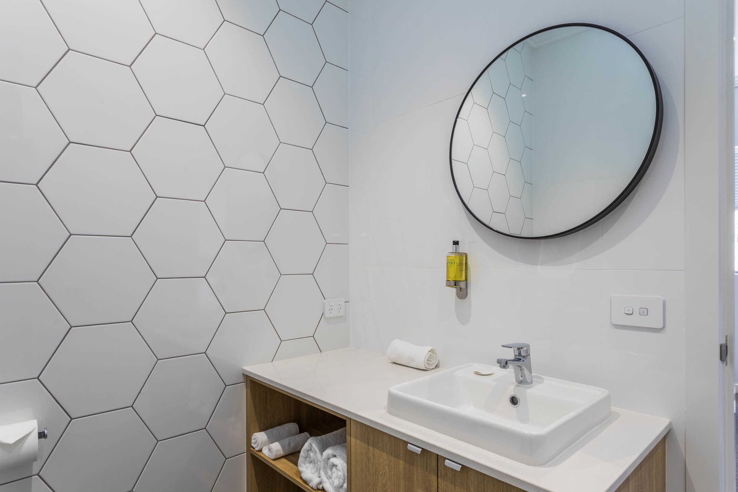 A bathroom with a hexagonal tile wall, a round mirror, a white sink, and a towel rack.