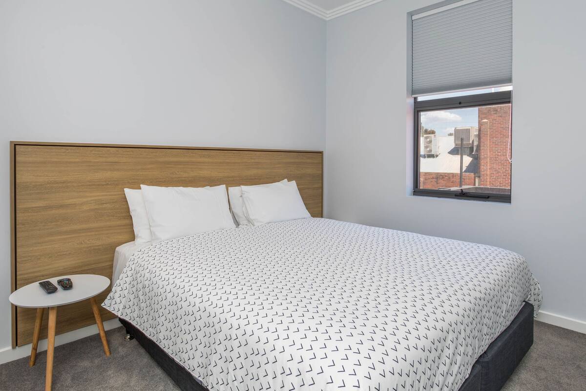 A bedroom with a wooden headboard, a white patterned bedspread, a small round table with a remote control, and a window with a view of buildings.