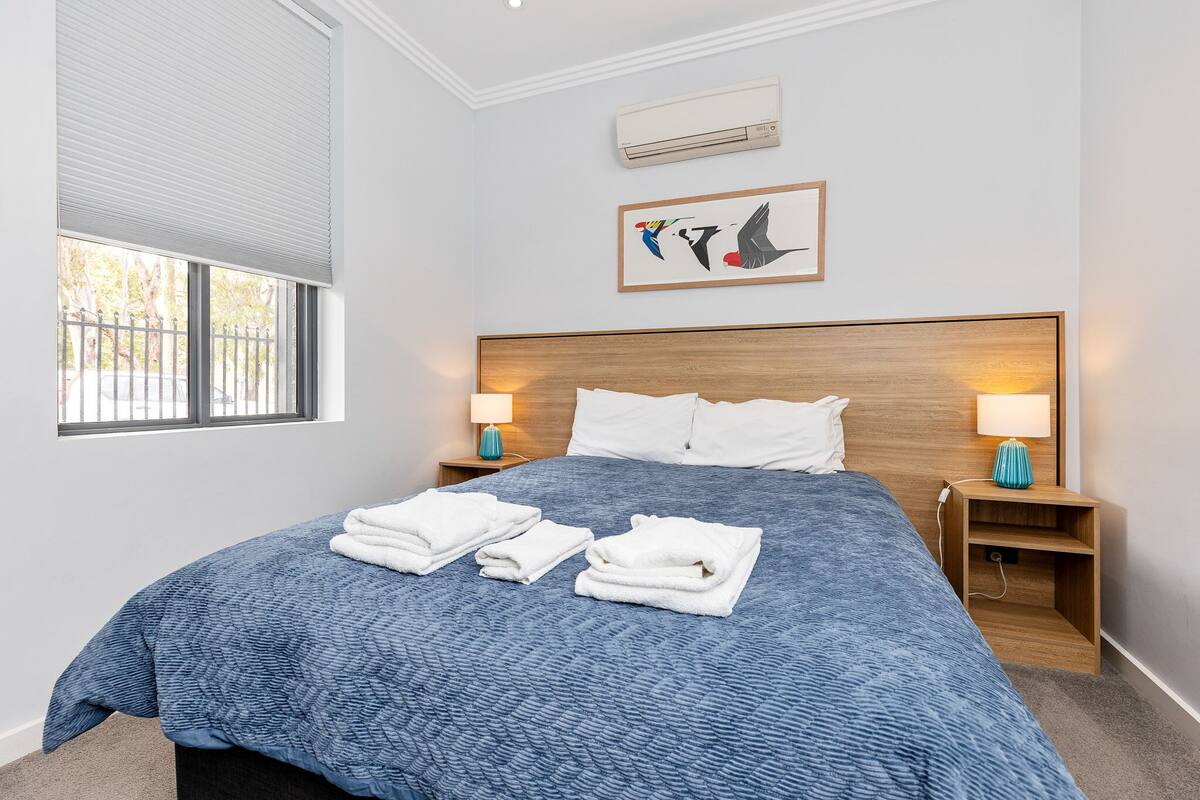 A hotel room with a large bed, two bedside tables with lamps, a window with blinds, and a wall-mounted air conditioner.