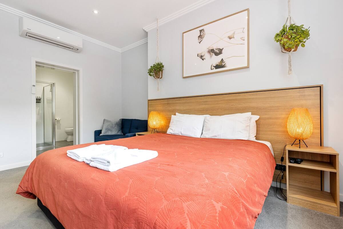 A modern bedroom with a large bed, two bedside lamps, a framed artwork, and a potted plant.