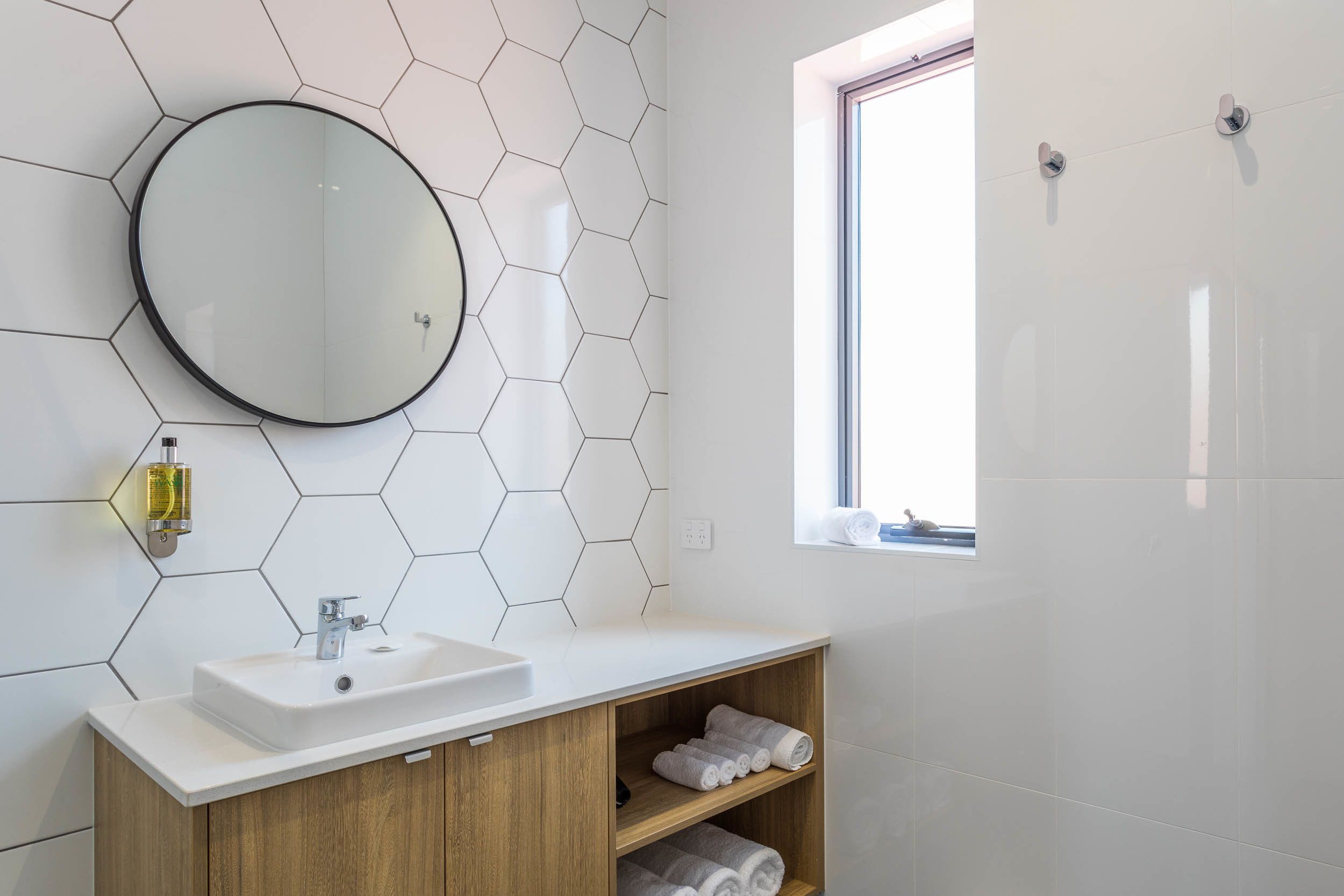 A modern bathroom with a round mirror, white hexagonal tiles, a white sink, wooden vanity, and a window with towels.