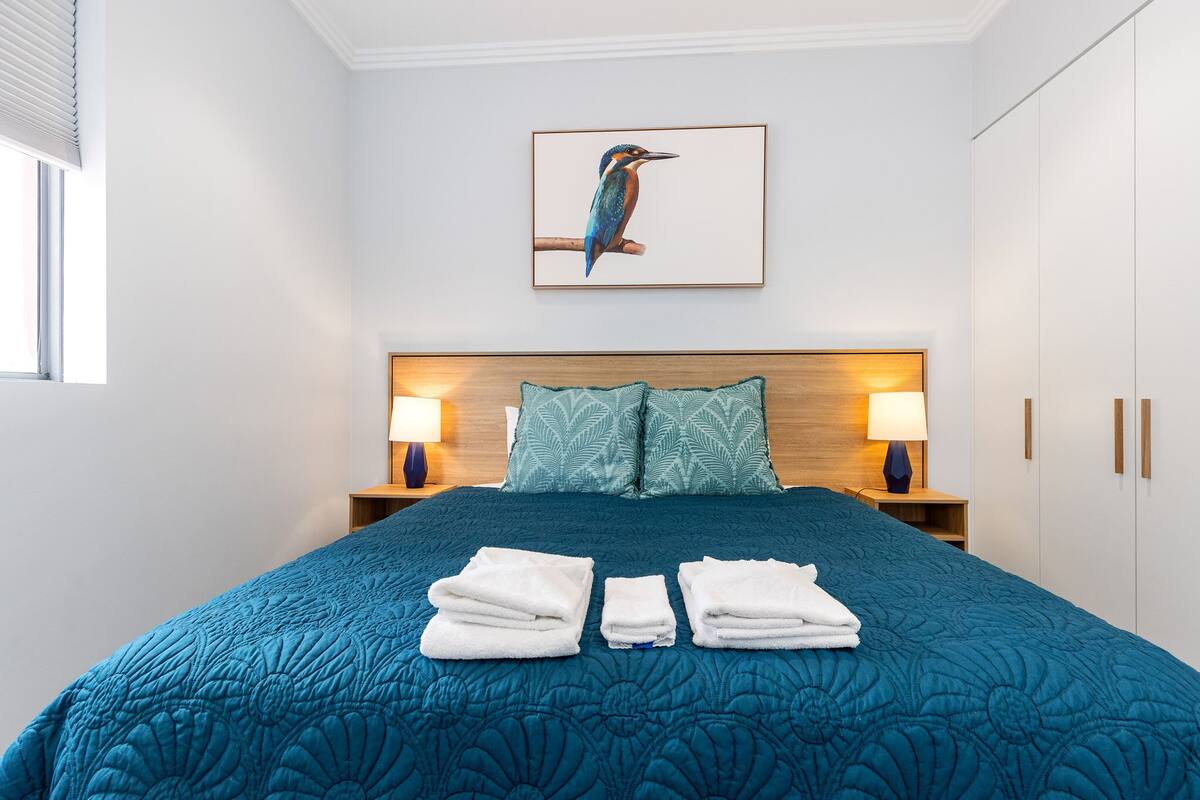 A neatly made bed with a blue quilt, two pillows, and three folded towels on the bed.