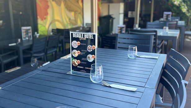 An outdoor dining area with a menu board for Beagle Hotel Specials, including items like $25 desserts and $25 drinks.