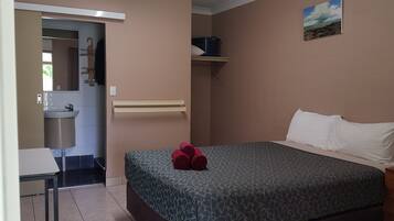 Une chambre à coucher comprenant un lit, une table de chevet, une commode, une étagère fixée au mur, et une salle de bain visible à travers une porte ouverte.